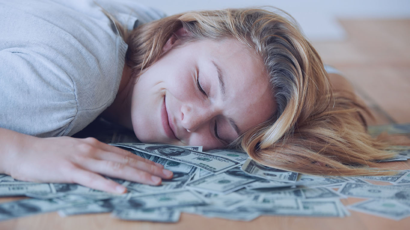 Escolher as dezenas das loterias: Uma menina sorridente com os olhos fechados, deitada no chão com a cabeça em uma pilha de dinheiro