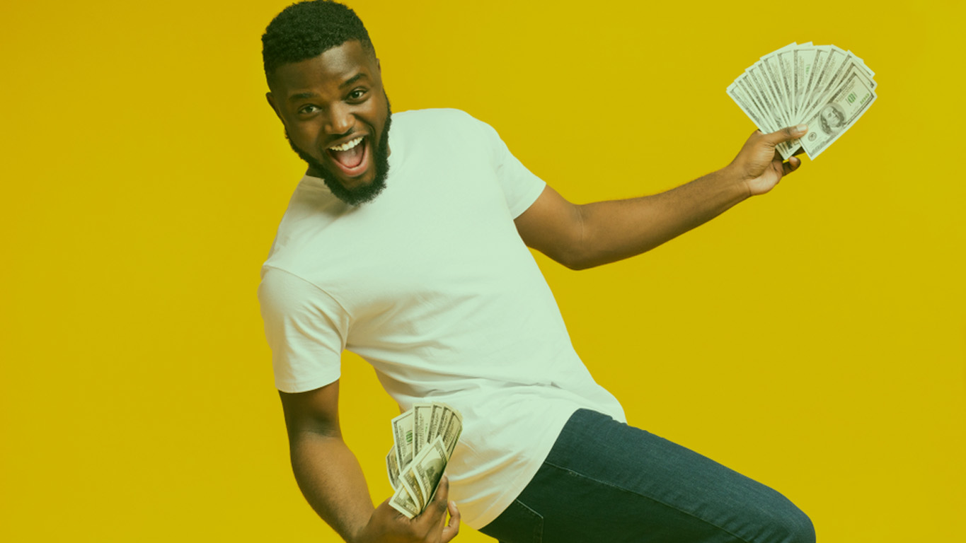homem posando para a foto em uma parede branca usando calça jeans, camiseta branca e sorrindo com muitas notas de dinheiro na mão