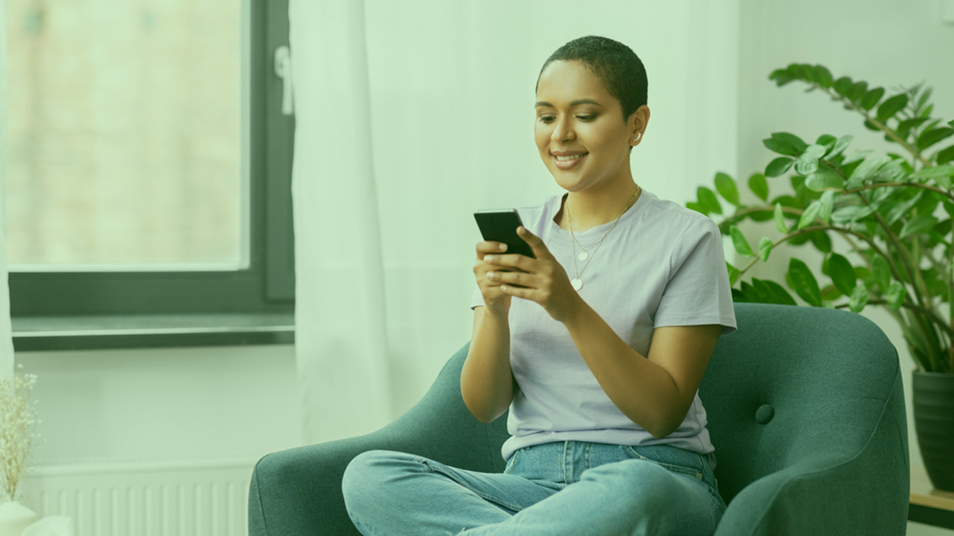 mulher careca sentada em uma poltrona usando calça jeans, camiseta lilás enquanto sorri olhando para o seu celular