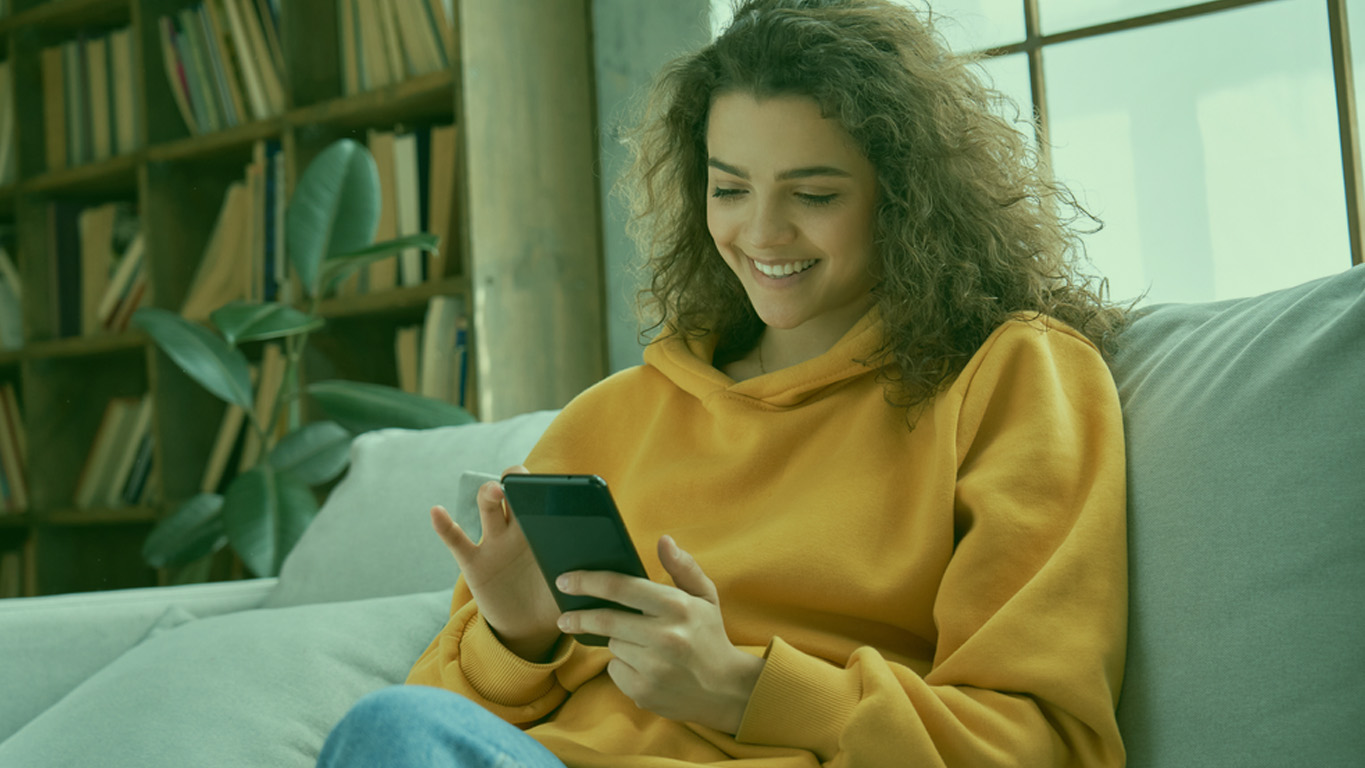 Aprenda Jogar na Mega-Sena Online: Mulher sentada no sofá sorrindo, vestindo blusa de frio amarela com uma planta ao lado e cabelo solto