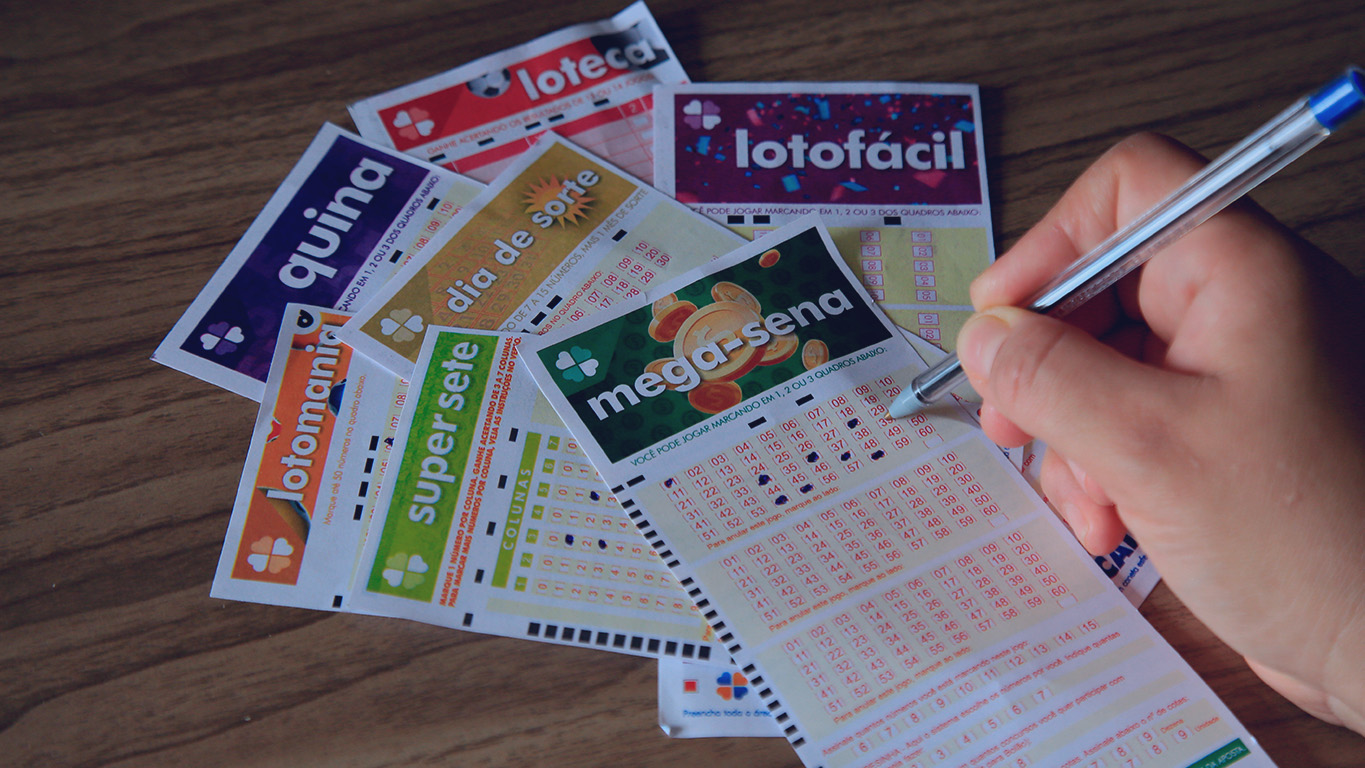 Mão marcando números em bilhetes de aposta das Loterias Caixa em mesa de madeira.