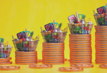 Cestas de compras com alimentos em cima de pilhas de moedas enfileiradas em fundo amarelo. Representa o aumento dos preços no Brasil.