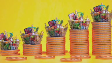 Cestas de compras com alimentos em cima de pilhas de moedas enfileiradas em fundo amarelo. Representa o aumento dos preços no Brasil.