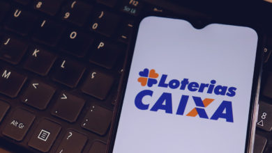 Logo da Loterias Caixa na tela do smartphone em cima de um teclado de laptop.