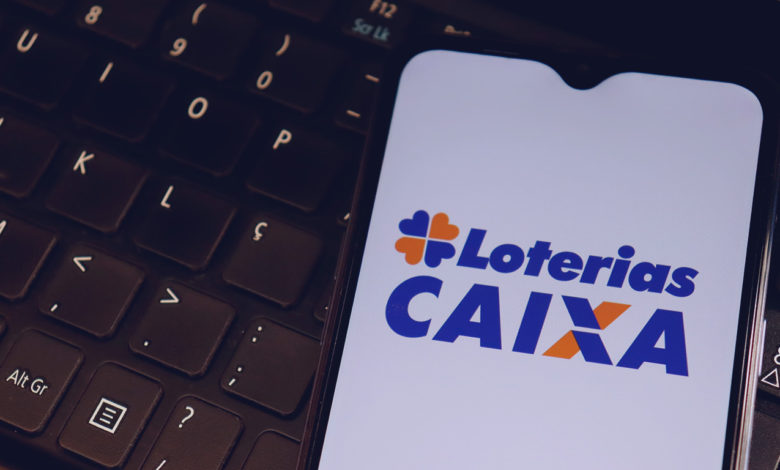 Logo da Loterias Caixa na tela do smartphone em cima de um teclado de laptop.