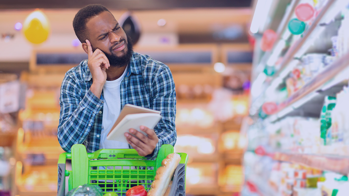 Homem negro comprando comida com lista de compras nas mãos, em pé com carrinho perto da prateleira no supermercado. Sua expressão fácil demonstra confusão e desapontamento com o preço dos produtos.
