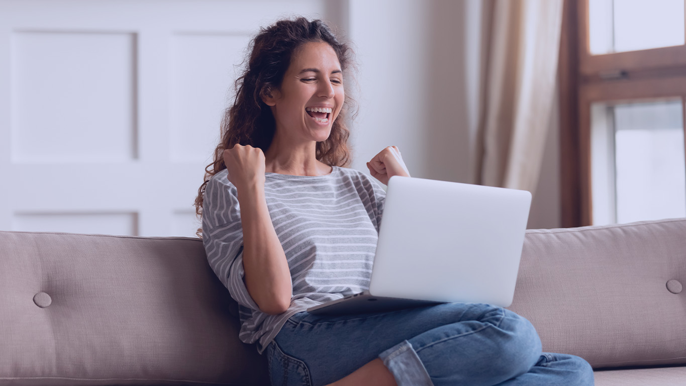 Mulher sentada no sofá com laptop no colo e punhos cerrados grita de alegria enquanto olha para a tela do computador. Provavelmente comemora vitória. Como ganhar no LottoCap YESSS.