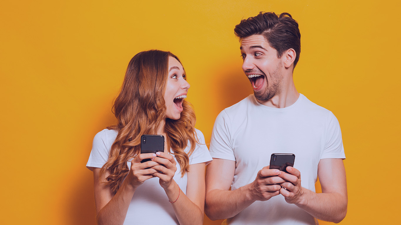 Um jovem homem e uma jovem mulher felizes, um olhando para o outro, cada um com um celular em mãos.